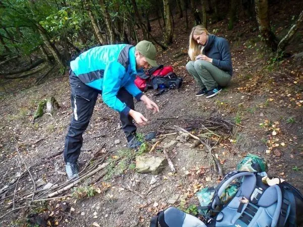 Mann sammelt Äste für ein Feuer, Frau sitzt mit Smartphone im Hintergrund