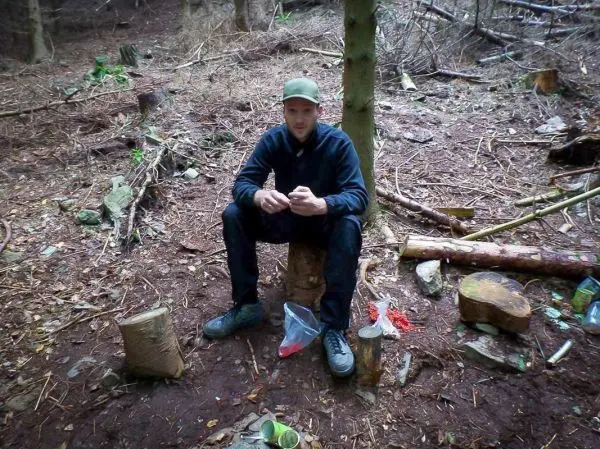 Mann sitzt auf einem Holzscheit im Wald, umgeben von Ästen und Holzstücken
