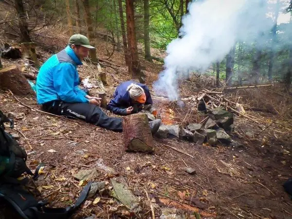 Person bläst in ein Feuer, während eine andere Person zuschaut