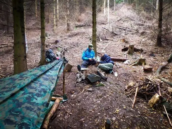 Tarp-Notunterkunft aus Camouflage-Material und zwei Personen am Boden mit Holz