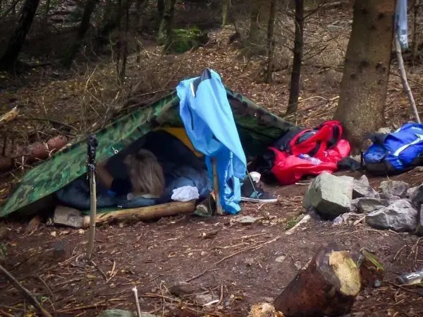 Tarp-Notunterkunft aus Camouflage-Plane mit Rucksack und Holzstücken im Hintergrund