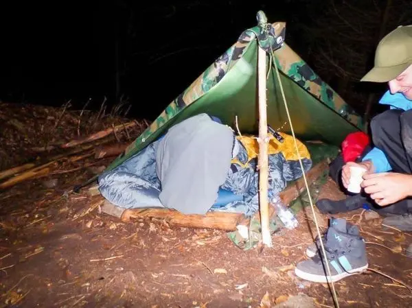 Tarp-Notunterkunft aus Camouflage-Plane mit Schlafsack und Person daneben, die eine Tasse hält