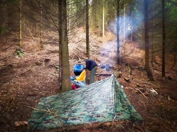 Tarp-Notunterkunft aus Tarnmaterial im Wald, Rauch steigt aus einem kleinen Feuer auf