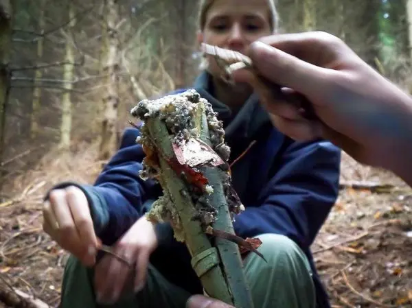 Zunder wird auf einen Holzstock gelegt, während eine Person im Hintergrund sitzt