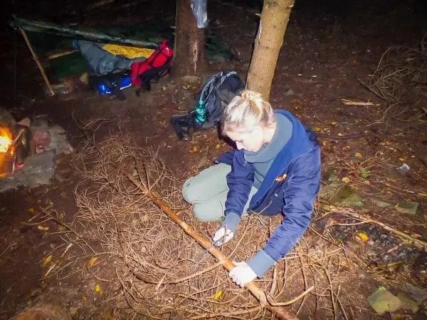 Zweig wird mit einem Messer bearbeitet, während ein kleines Feuer brennt