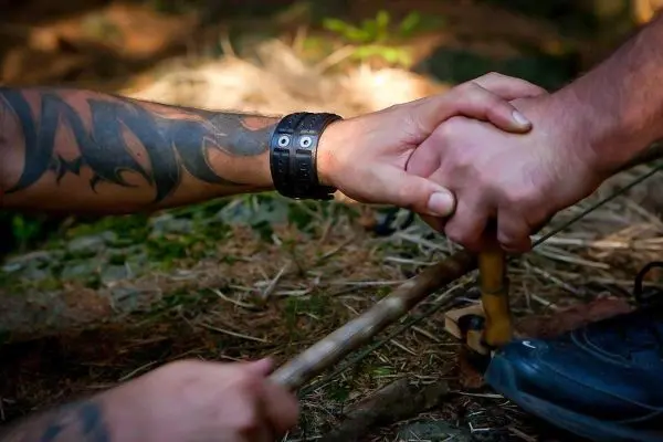 Hände greifen sich gegenseitig, während ein Holzstück für Feuerbohrer vorbereitet wird