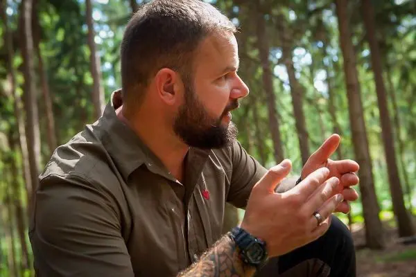 Mann gestikuliert im Wald, umgeben von hohen Bäumen