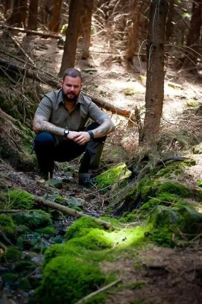 Mann hockt in einem Waldgebiet neben einem kleinen Bachlauf auf moosbedecktem Boden