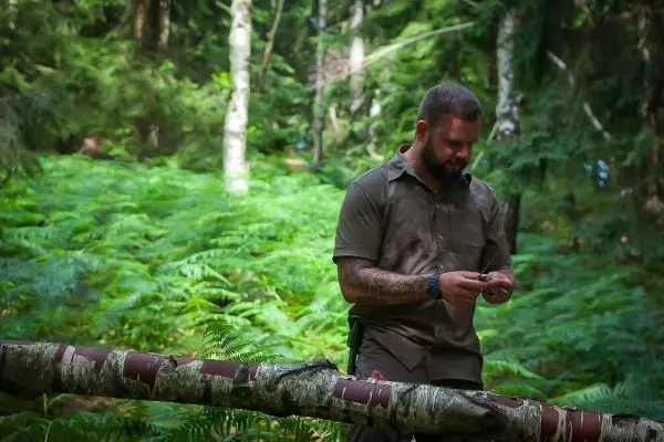 Mann prüft Ausrüstung auf einem Baumstamm im Wald zwischen Farnen