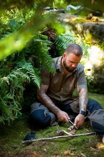 Mann schnitzt mit einem Messer an einem Holzstück im Wald