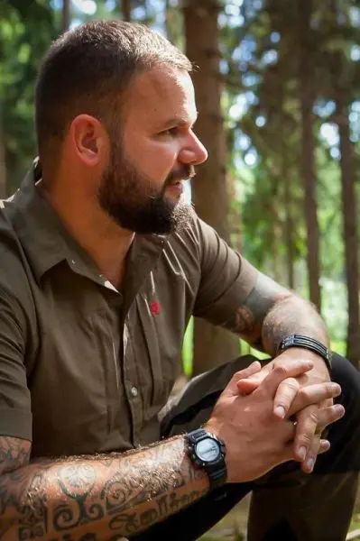 Mann sitzt im Wald, Hände gefaltet, mit Tätowierungen und Armbanduhr