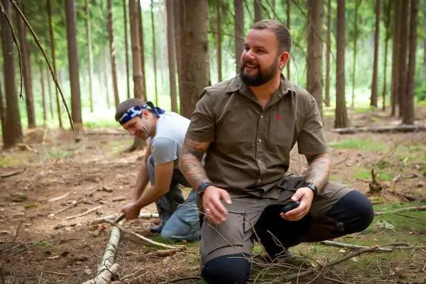 Zwei Personen arbeiten im Wald, einer kniet und hält ein Werkzeug, der andere bearbeitet einen Stock