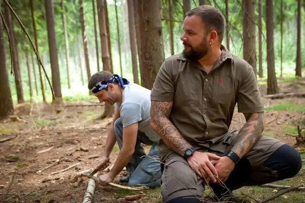 Zwei Personen arbeiten im Wald, einer schnitzt mit einem Messer an einem Stock