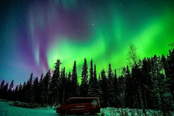 Bunte Nordlichter leuchten über schneebedeckten Bäumen und einem parkenden Fahrzeug