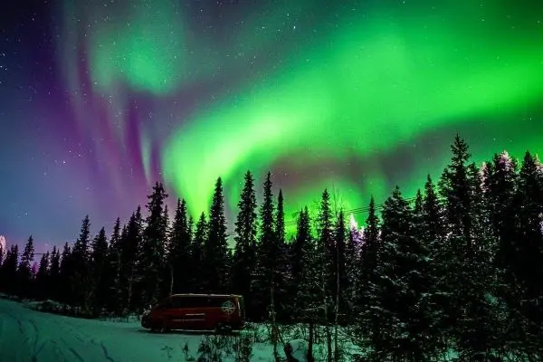 Grüne Polarlichter leuchten über schneebedeckten Bäumen und einem Fahrzeug