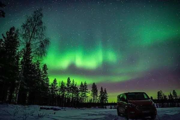 Grünes Polarlicht leuchtet über schneebedecktem Boden und Bäumen, Fahrzeug im Vordergrund
