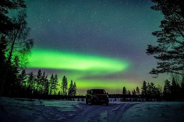 Grünes Polarlicht leuchtet über verschneiter Landschaft mit Bäumen und einem Fahrzeug
