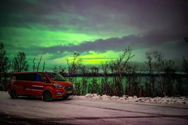 Orangefarbener Van steht auf verschneiter Straße vor einem See unter Polarlichtern
