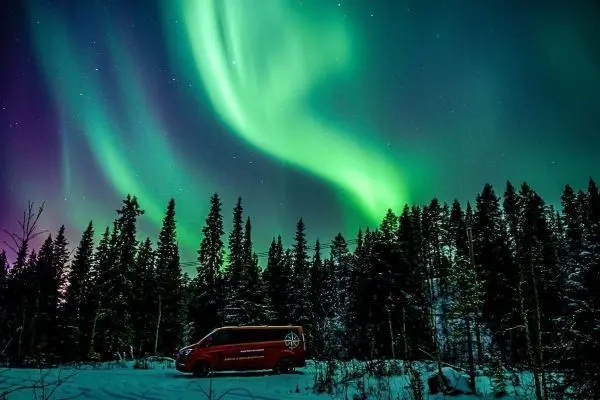 Rote Fahrzeug steht im Schnee vor einem Wald unter dem Polarlicht
