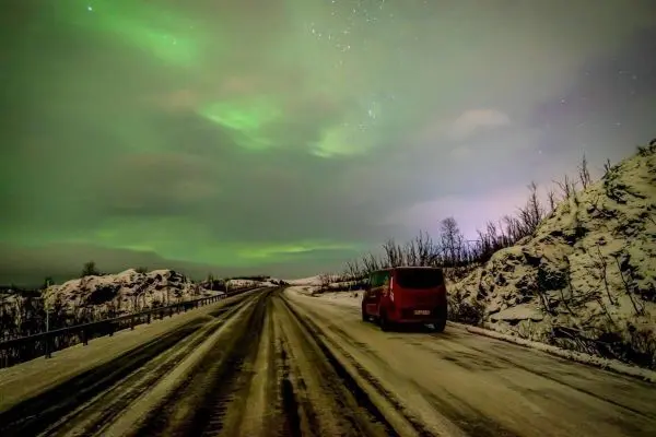 Rotes Fahrzeug steht auf einer verschneiten Straße unter Polarlichtern