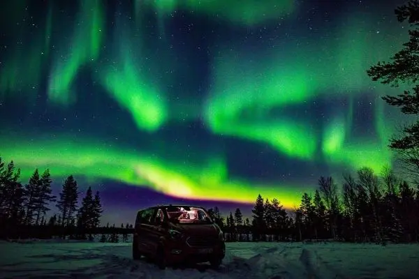 Sternenhimmel mit Polarlichtern über einem Fahrzeug im Schnee und Bäumen
