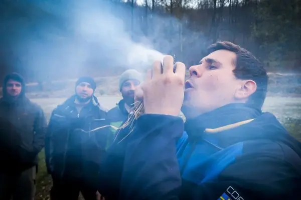 Mann bläst in Zunder, Rauch steigt auf, mehrere Personen beobachten