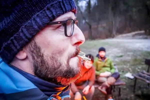 Mann mit Brille und Mütze sitzt am Lagerfeuer, im Hintergrund sind weitere Personen