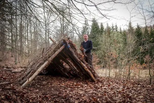 Mann steht neben einer selbstgebauten Tarp-Notunterkunft aus Ästen und Laub