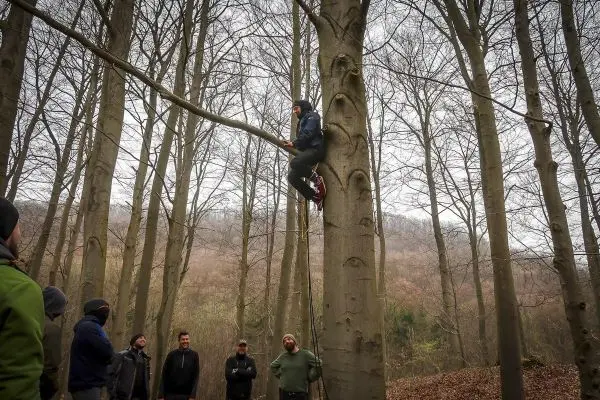 Person klettert an einem Baumstamm hoch, Gruppe schaut zu beim Outdoorkurs
