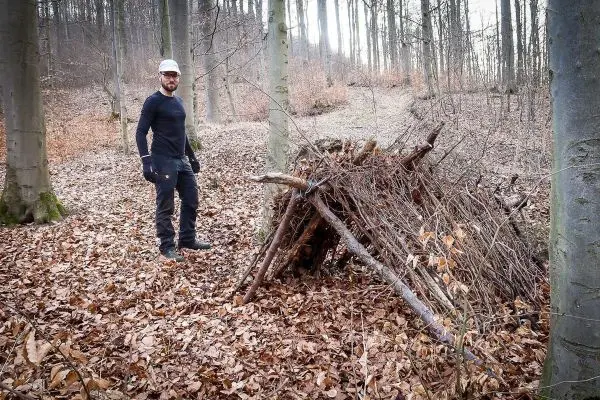 Person steht neben einer selbstgebauten Unterkunft aus Ästen