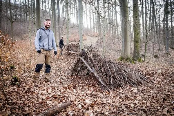 Selbstgebaute Laubhütte aus Ästen im Wald, zwei Personen im Hintergrund