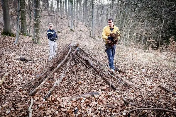 Zwei Personen stehen neben einer selbstgebauten Laubhütte im Wald