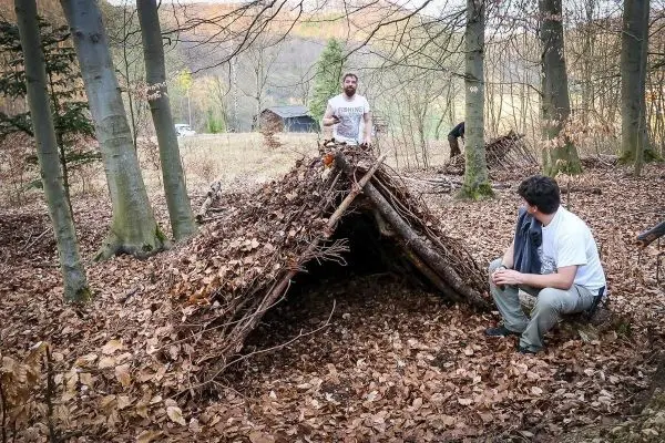 Zwei Personen stehen und sitzen neben einer selbstgebauten Tarp-Notunterkunft aus Ästen und Laub