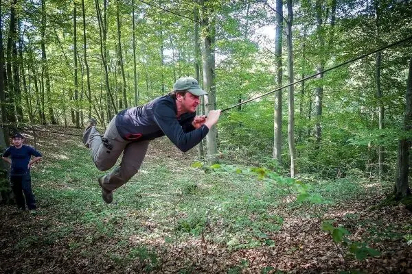 Mann überquert selbstgebaute Seilbrücke im Wald, während ein Zuschauer zusieht
