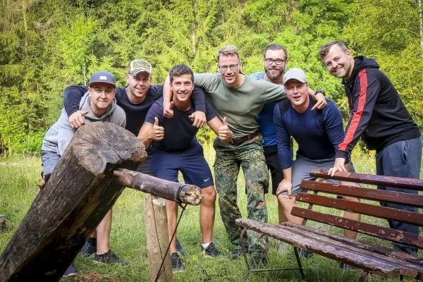 Sechs Personen posieren lächelnd vor einer Holzbank und einem Holzstück im Freien