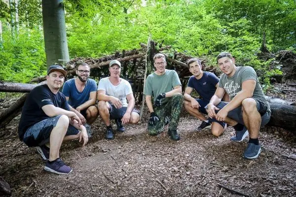 Sechs Personen posieren vor einer selbstgebauten Holzunterkunft im Wald