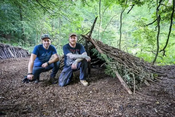 Zwei Personen knien vor einer selbstgebauten Laubhütte aus Ästen und Zweigen
