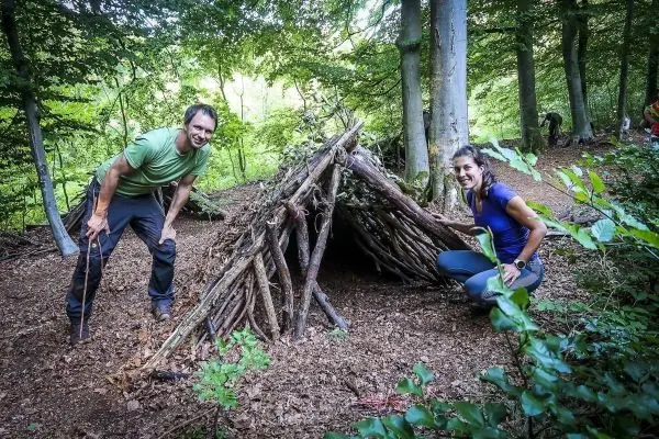 Zwei Personen stehen neben einer selbstgebauten Laubhütte aus Ästen und Zweigen