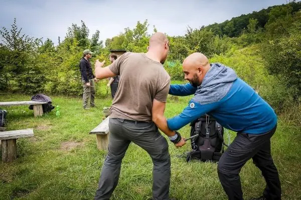 Zwei Personen üben eine Technik im Freien, im Hintergrund stehen weitere Personen