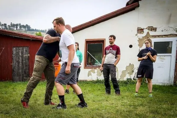 Zwei Personen üben einen Nahkampfgriff auf einer Wiese vor einem Gebäude