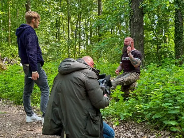 Drei Personen im Wald, eine sitzt auf dem Boden und spricht, zwei stehen daneben