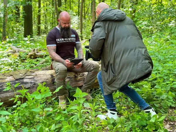 Zwei Personen sitzen und stehen auf einem Baumstamm im Wald, eine Person hält ein Tablet