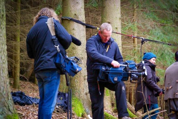 Drei Personen im Wald tragen Kamera- und Audioausrüstung zwischen Bäumen