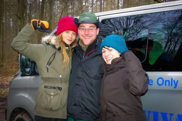 Drei Personen posieren lächelnd vor einem Fahrzeug im Wald