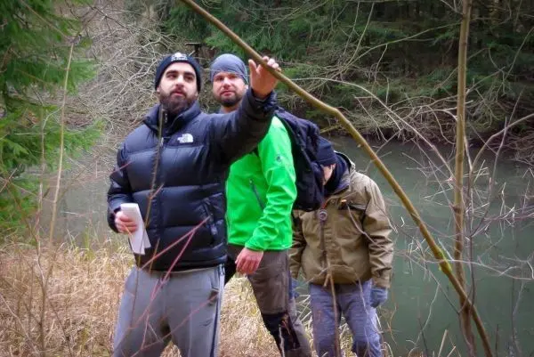 Drei Personen stehen am Ufer eines Gewässers und betrachten einen Baumzweig