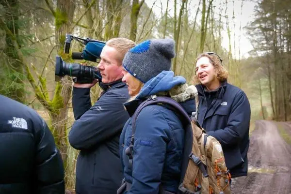 Drei Personen stehen auf einem Waldweg, eine filmt mit einer Kamera