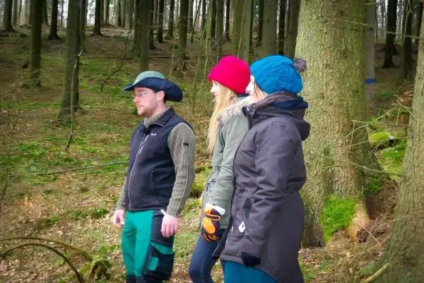 Drei Personen stehen im Wald zwischen Bäumen und betrachten die Umgebung