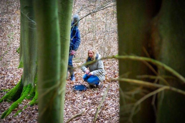 Frau kniet im Laub und untersucht einen Gegenstand in einer Waldlandschaft