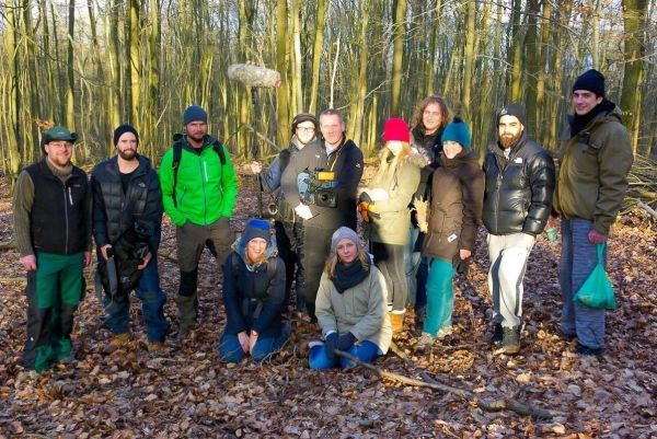 Gruppe von Personen steht im Wald auf Laubboden, einige halten Ausrüstung
