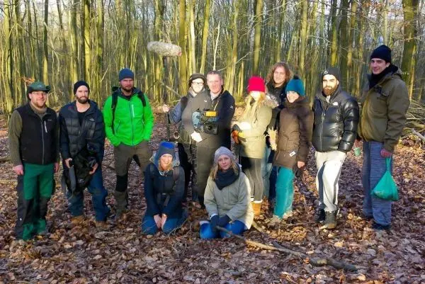 Gruppe von Personen steht im Wald auf Laubboden, einige halten Ausrüstung